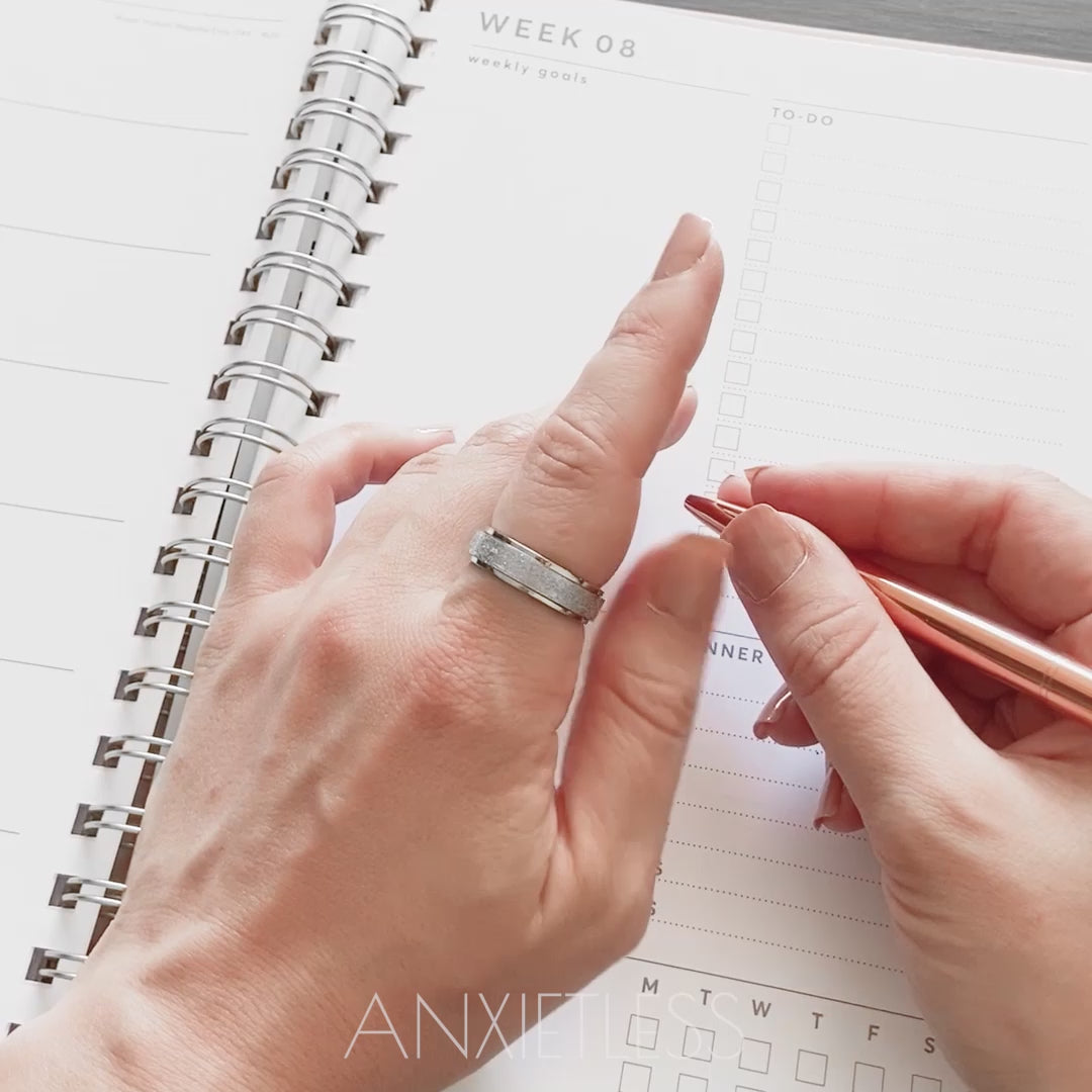 Woman demonstrating spinning action of silver anxiety ring while holding rose gold pen with other hand, with diary in background. Close-up of spinning silver and rose gold anxiety rings on thumb in cityscape