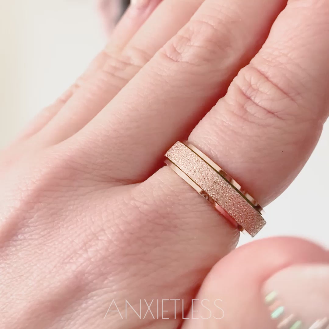Woman spinning rose gold anxiety ring on index finger in close-up, then moving hand further away with grey and pink wall decoration in the background. Second half of video shows spinning rose gold anxiety ring with city buildings in the background