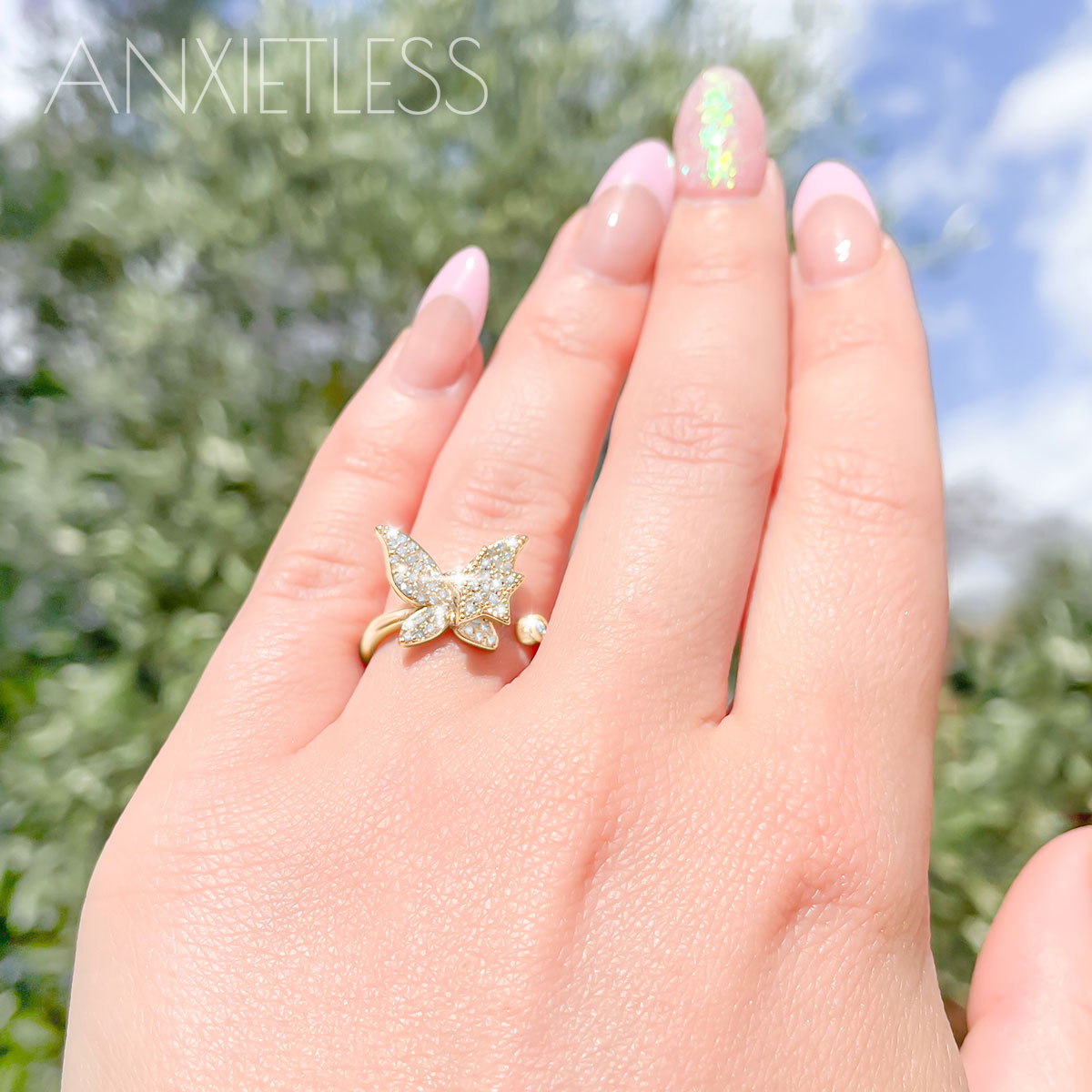 Woman's hand showcasing gold butterfly anxiety ring with star charm, against a backdrop of trees and sky