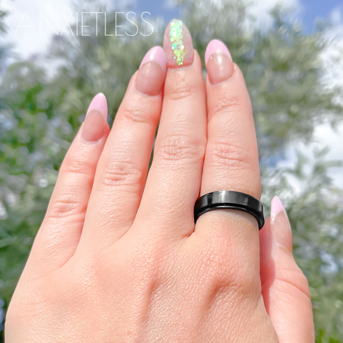 Woman with pink nails wearing a black fidget ring, featuring a smooth polished design, with blurry trees and sky in the background