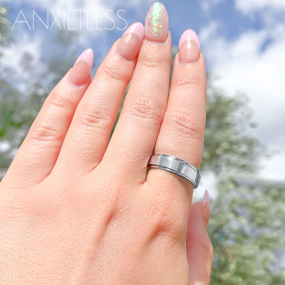 Girl with pink nails wearing a silver fidget ring, featuring a smooth polished design, with blurry trees and sky in the background