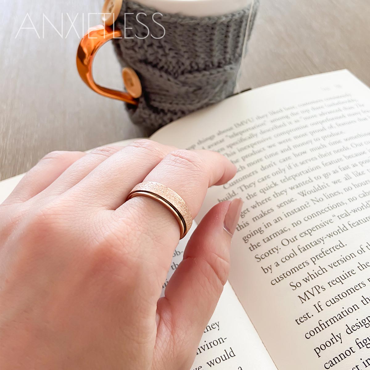 Woman wearing rose gold anxiety ring on index finger, with a book and a mug with a grey knitted cover in the background