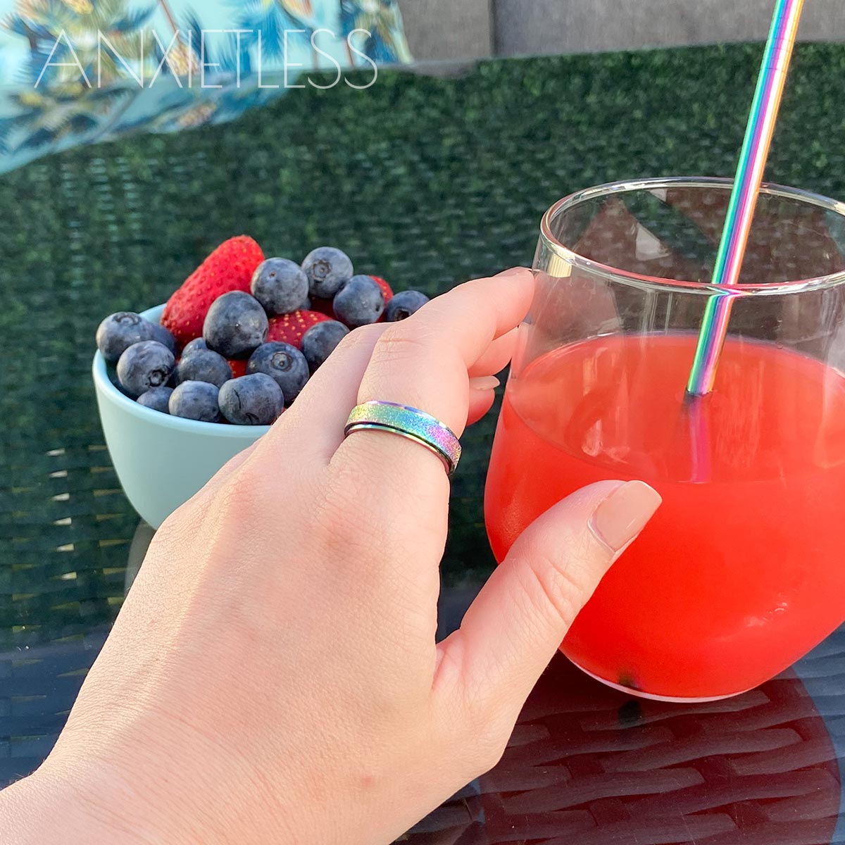Woman wearing rainbow anxiety ring on index finger, holding a glass with red juice and a rainbow straw. In the background, there is a blue bowl with strawberries and blueberries, a glass table, and a grey couch with a blue palm leaf pillow.